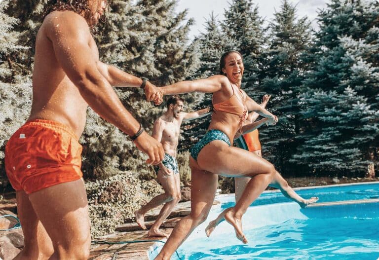 Woman and friends jumping into a backyard pool, smiling as they enjoy a sunny day surrounded by trees.